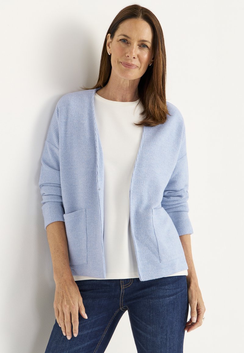 Vaaleansininen ribattu neuletakki, jossa on avointa etuosaa ja kaksi taskua, kerrottu valkoisen paidan päällä. Yhdistetty tummiin farkkuihin, jotka ovat sileäpintaiset.