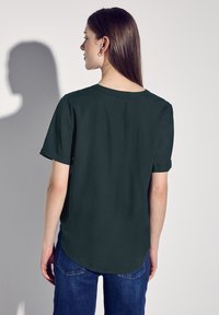 Camicetta verde scuro a maniche corte con vestibilità rilassata, scollatura rotonda e maniche arrotolate. Tessuto morbido con orlo dritto e taglio alto-basso.