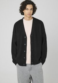 Cardigan noir à côtes avec un décolleté en V profond, six boutons sur le devant et une coupe décontractée, porté par-dessus une chemise rose pâle.