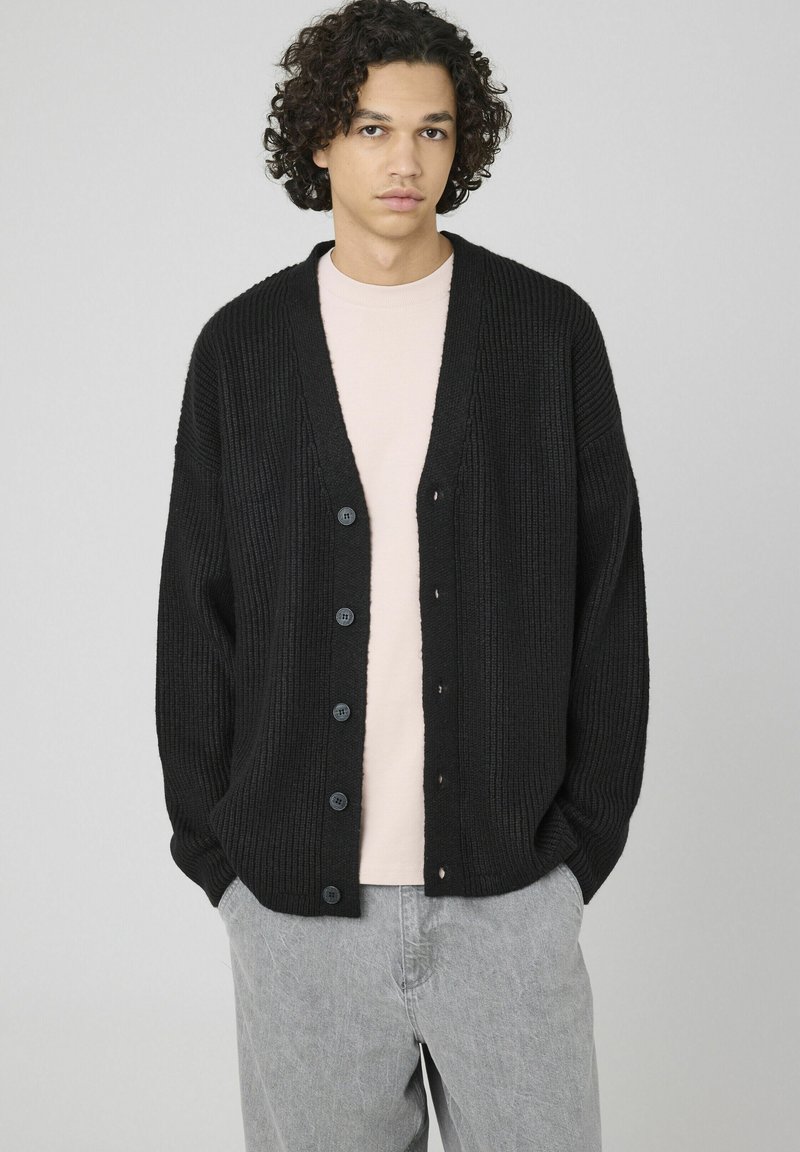 Cardigan noir à côtes avec un décolleté en V profond, six boutons sur le devant et une coupe décontractée, porté par-dessus une chemise rose pâle.