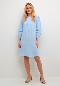 Ljust blå klänning med krage, kort knappslå och puffärmar med spetsdetaljer. Modell som bär beiga klackskor med blockklack.