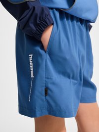 Niebieskie krótkie spodenki sportowe z boczną kieszenią, włożoną ręką i widocznym granatowym elastycznym rękawem kurtki.