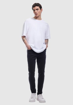 Biała bawełniana koszulka o luźnym kroju, w zestawie z czarnymi jeansami slim fit i białymi trampkami. Widoczne detale skóry przez tatuaże.