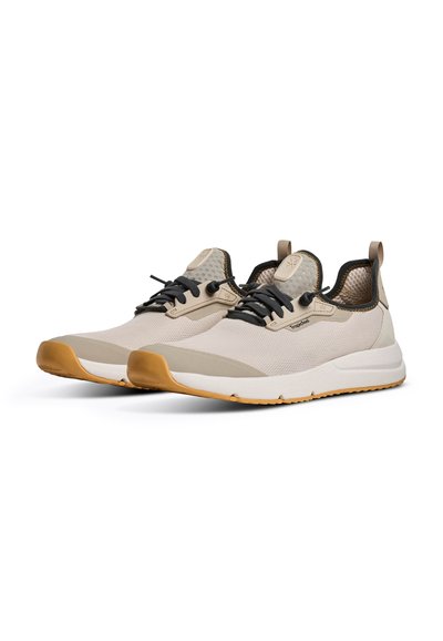 Paire de chaussures de sport beige et blanche avec lacets noirs, semelles en caoutchouc marron clair et tige en tissu mesh, positionnées à un léger angle sur fond blanc.