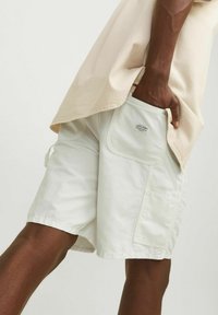 Pantaloni cargo bianchi in cotone con una tasca laterale e un'etichetta. Abbinate a una camicia beige, morbida e ampia. La texture sembra morbida e leggera.