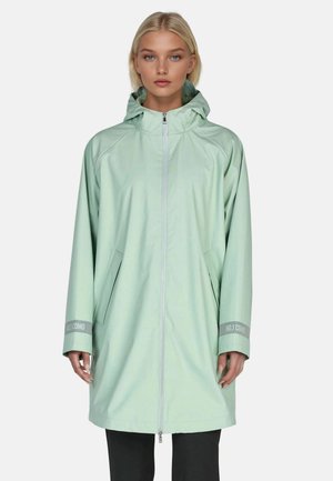 Blonde woman wearing a light green hooded raincoat zipped up over black pants, standing against a plain background.