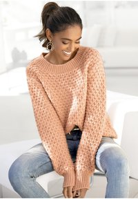 Helles Pfirsichfarbenes Strickoberteil mit einem strukturierten Rautenmuster, weiter Rundhalsausschnitt und gerippten Bündchen. Kombiniert mit blauen Jeans.