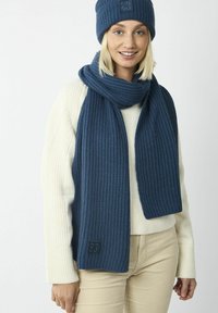 Marineblaue gestrickte Mütze und Schal-Set mit gerippter Textur und Aufnäher. Der Schal hängt elegant und ergänzt einen cremefarbenen Pullover.