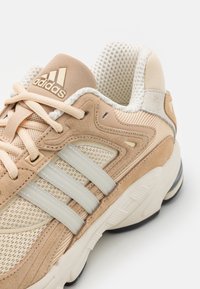 Baskets de sport beiges en matériaux mesh et suédé, avec trois rayures blanches, une semelle rembourrée et un col de couleur crème.