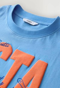 T-shirt en coton bleu clair avec un col rond. Présente de grandes lettres orange et une broderie bleue sur le devant. Étiquette du fabricant visible.