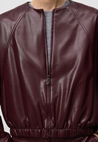 Giacca in pelle bordeaux con collo rotondo, chiusura a zip e cintura elastica; texture liscia e design aderente con maniche a raglan.