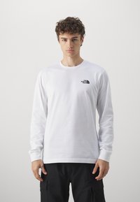Hvid langærmet t-shirt lavet af bomuld. Har et lille sort logo på venstre bryst og en rund halsudskæring. Bæres med sorte cargo-bukser.
