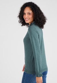 Groen gebreid trui met een ruches-kraag, textuur met bobbelontwerp en geribde manchetten. Gecombineerd met blauwe jeans, zijaanzicht weergegeven.