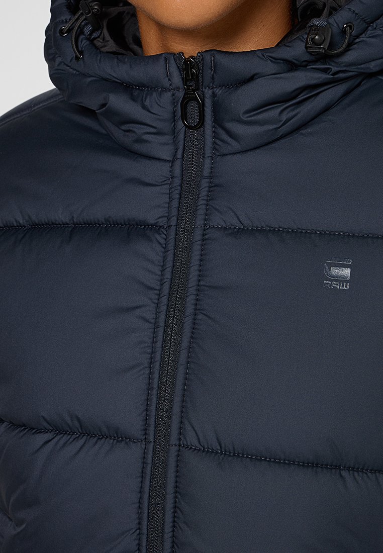 Marineblauer Puffer-Jacke mit gequiltetem Design, ausgestattet mit einem hohen Kragen, einem Reißverschluss vorne und einem kleinen Logo auf der Brust. Weiche, glatte Textur.