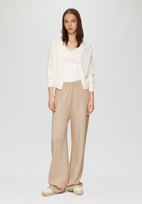 Witte cardigan over een witte geribde top, gecombineerd met beige wijde broek. Getextureerde stof en een aansluitende tailleband. Casual espadrille schoenen.