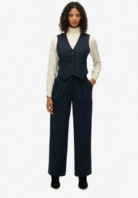 Gilet a righe navy con cinque bottoni, abbinato a pantaloni a gamba larga e un dolcevita crema. Texture liscia con una vestibilità su misura.
