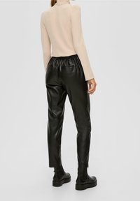 Schwarze Kunstlederhose mit elastischem Bund, schmal geschnitten, und zwei Gesäßtaschen. Kombiniert mit einem beigen Rollkragenpullover.