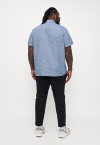 Camisa de botones de manga corta, color azul claro, en un tejido texturizado, combinada con pantalones negros y zapatillas deportivas. Costuras de hombro y yugo trasero visibles.