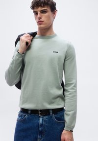 Νέος άνδρας που φοράει ανοιχτοπράσινο πουλόβερ BOSS και μπλε τζιν, κρατώντας ένα μαύρο μπουφάν στον ώμο του, σε ένα απλό λευκό φόντο.