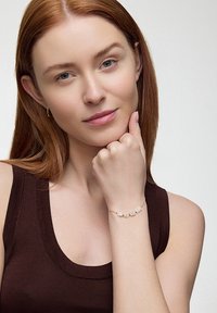 Złota bransoletka z przezroczystymi, prostokątnymi koralikami, noszona na nadgarstku. Modelka ma proste, czerwone włosy i nosi brązowy top na ramiączkach.