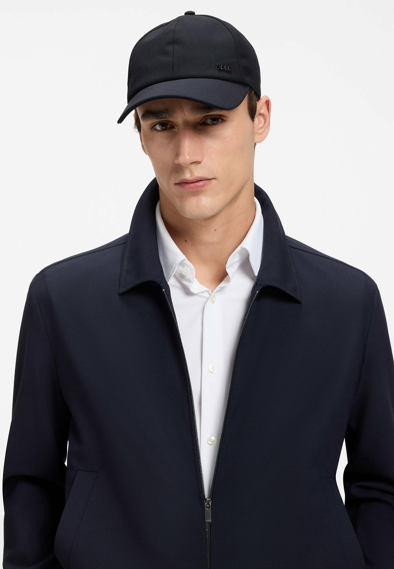 Jeune homme portant une casquette de baseball noire, une veste zippée bleu marine et une chemise blanche boutonnée sur un fond uni.