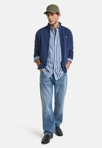 Marineblaue Reißverschlussjacke, hellblau-weiß gestreiftes Hemd, hellblaue Jeans, schwarze Schuhe, olivgrüne Kappe. Legere Kleidung mit strukturierter Schichtung.