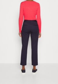 Osoba oblečená v jasně růžovém svetru s dlouhým rukávem, tmavě modrých kalhotách s vysokým pasem a tmavých teniskách, stojící zády k fotoaparátu na jednobarevném pozadí.