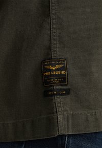 Dunkelolivgrüner Stoff mit strukturiertem Finish, versehen mit einem schwarzen und goldenen Etikett, auf dem "PME Legend", "American Classic" und "Typ Nr.: C-46" steht.