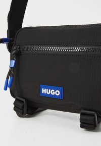 Crna torba za crossbody napravljen od teksturirane tkanine, s gornjim zatvaračem, plavim logom i podesivim remenom s čvrstim hardverom.