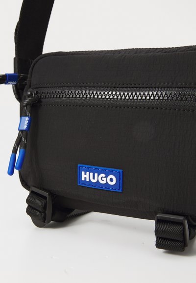 Черна чанта тип crossbody, изработена от текстурирана материя, с горно закопчаване с цип, синя логотипна нашивка и регулируемо рамо с здрави елементи.