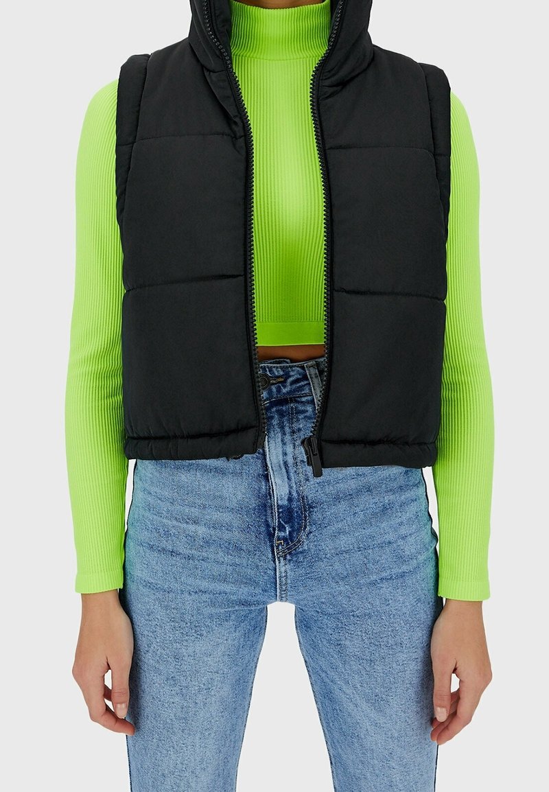 Personne portant un crop top côtelé à manches longues vert néon sous une doudoune noire et un jean bleu clair taille haute, les mains le long du corps.