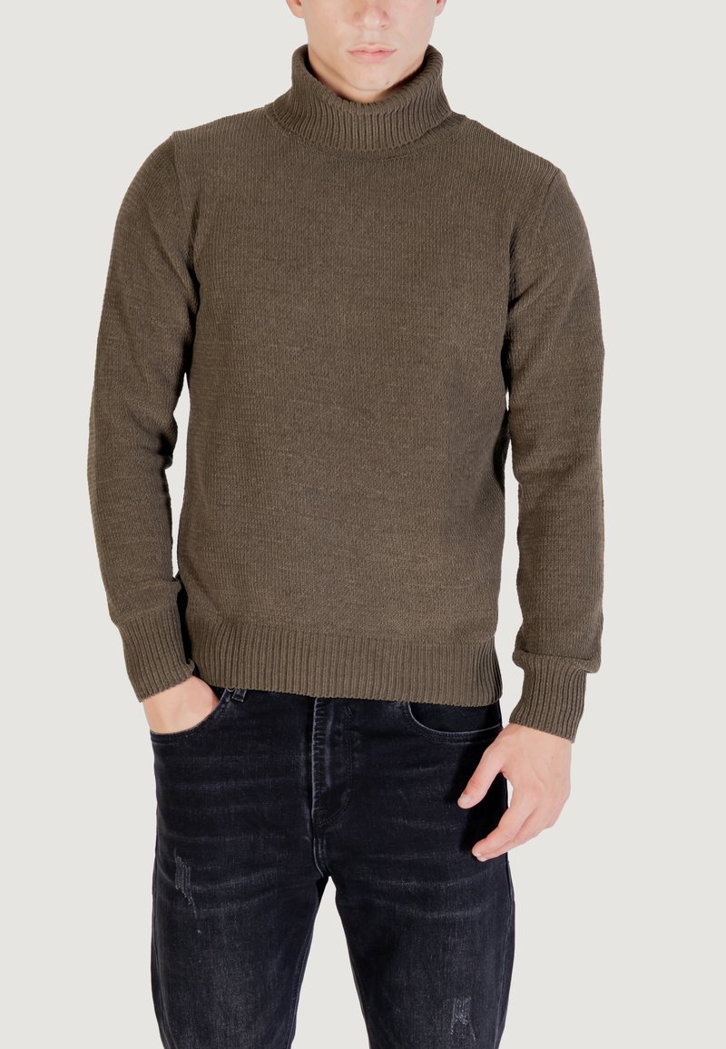 Uomo che indossa un maglione a collo alto a coste marrone con maniche lunghe e jeans neri sbiaditi, con una mano in tasca e uno sfondo neutro.