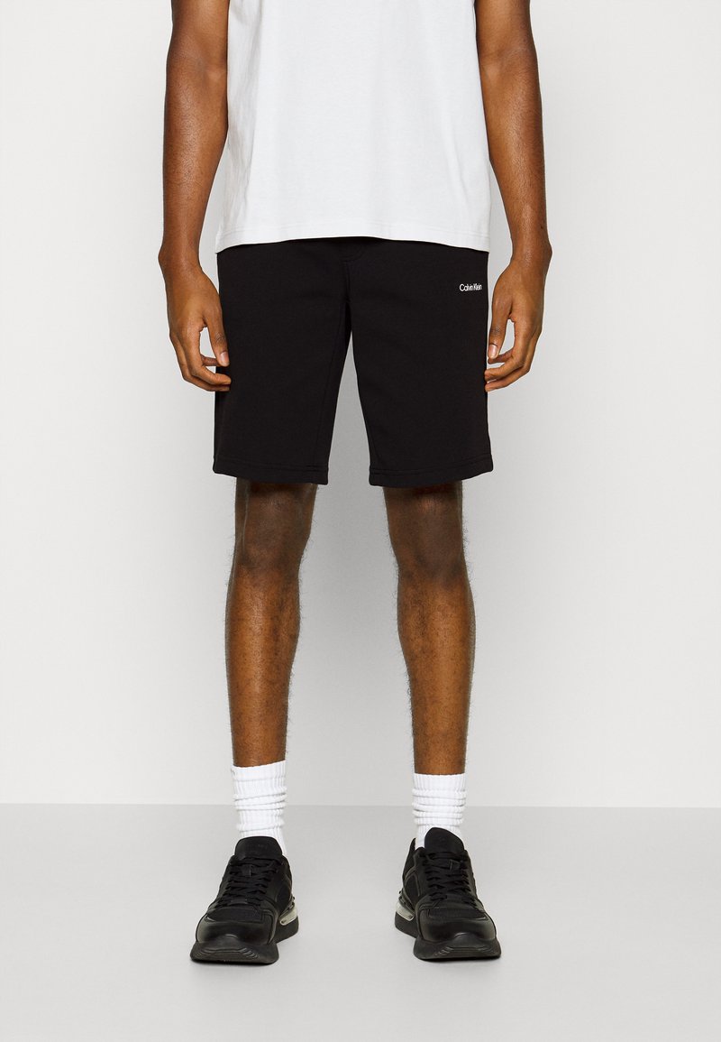 Sorte bomullsshorts med mellomlang lengde, med en liten hvit logo foran, kombinert med sorte sneakers og hvite sokker.