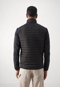 Sort isoleret jakke med en quiltet ryg og glatte ærmer, der har en høj krave og en tætsiddende silhuet. Stoffet fremstår struktureret og moderne.