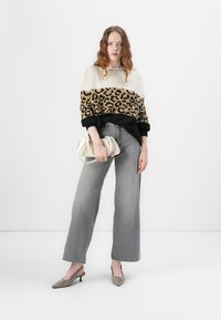 Een beige en zwarte sweater met luipaardprint en een blokkleurontwerp, gecombineerd met grijze wijde jeans en beige hakken, terwijl ze een witte tas vasthoudt.