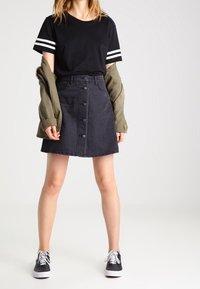 Camiseta negra de manga corta con rayas blancas, combinada con una falda de botones de denim negra y una chaqueta verde, complementada con zapatillas negras.