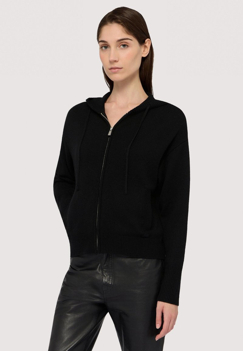 Felpa nera con zip realizzata in tessuto a maglia, dotata di cappuccio, lacci e polsini a costine. Abbinata a pantaloni in pelle nera.