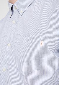 Primer plano de una camisa de botones a rayas azul y blanca con un bolsillo y una pequeña etiqueta roja HKT en el pecho.