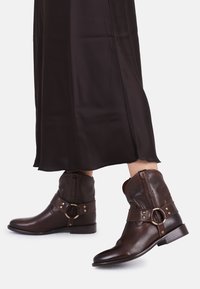 Donkerbruine leren enkellaarsjes met gouden ring- en banddetails, gedragen met een lange, donkerbruine rok op een witte achtergrond.