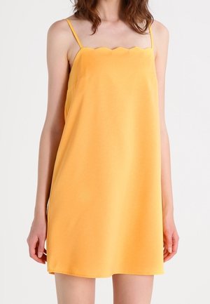 Kvinde iført en gul kjole uden ærmer med tynde stropper og en bølget halsudskæring, stående foran en ensfarvet hvid baggrund.