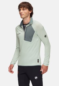 Light green pullover with a gray chest panel, zip pocket, and textured fabric. Features a high collar and a black logo on the sleeve.