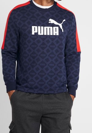 Mężczyzna w granatowej bluzie Puma z czerwonymi paskami na ramionach i ciemnoszarych spodniach, z rękami w kieszeniach, stojący na jednolitym tle.