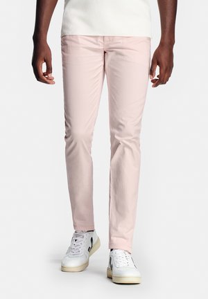 Man die lichtroze slanke broek en witte sneakers met zwarte accenten draagt, staand tegen een effen witte achtergrond.