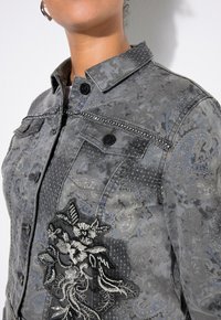 Grå denimjacka med embossade blommönster, silverpärlor och svart blommigt broderi på framsidan. Har en klassisk krage och knappar.