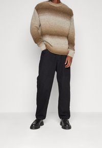 Sweter z dzianiny o okrągłym dekolcie w gradientowej brązowej kolorystyce, z teksturowanej tkaniny, zestawiony z luźnymi czarnymi spodniami i czarnymi butami.