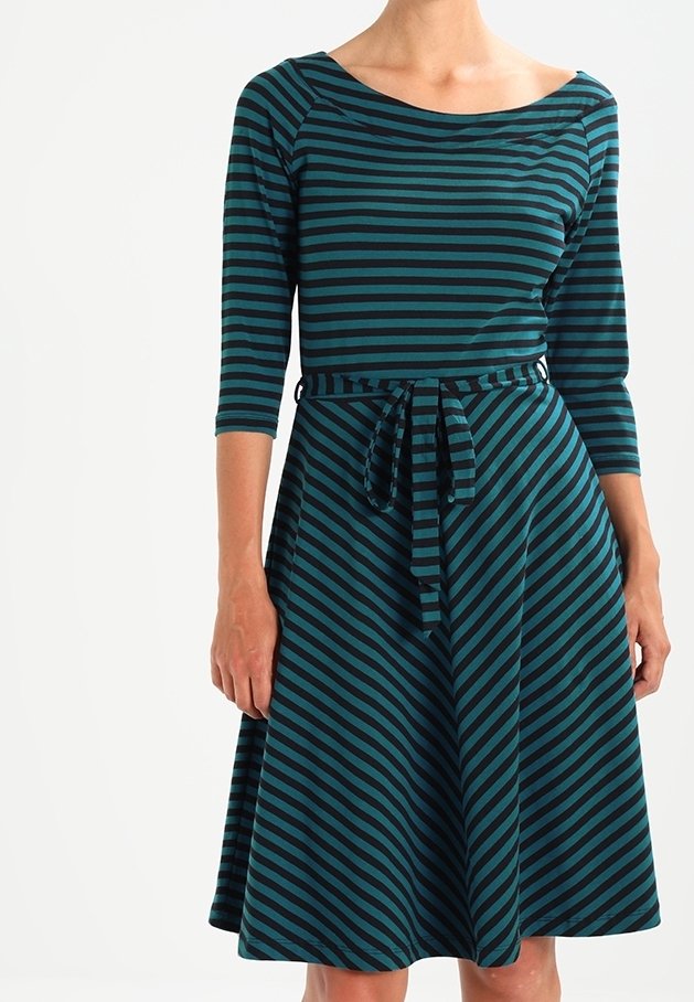 Femme portant une robe rayée bleu-vert et noire à manches trois-quarts avec une ceinture nouée à la taille, debout devant un fond blanc.
