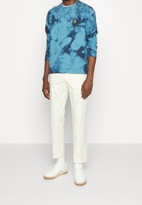 Blue tie-dye sweatshirt with a round neckline and a black logo patch, paired with white chinos and white sneakers. Smooth fabric texture.