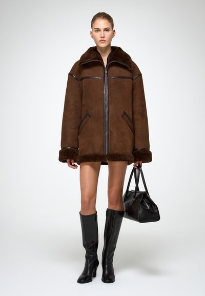 Pruun nahksoonega ülisuur jope, millel on karusnahast ääris, lukuga sulgemine ja suured taskud; kombineeritud mustade põlvini ulatuvate saapade ja musta käekotiga.