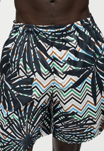 Gros plan d'une personne portant un short multicolore à motif zigzag avec de grandes feuilles tropicales noires imprimées et une taille élastique.