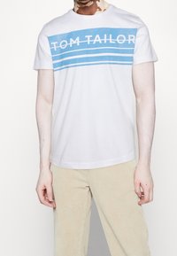 T-shirt en coton blanc avec des rayures horizontales bleues et le texte "TOM TAILOR" en blanc. Manches courtes et col rond.